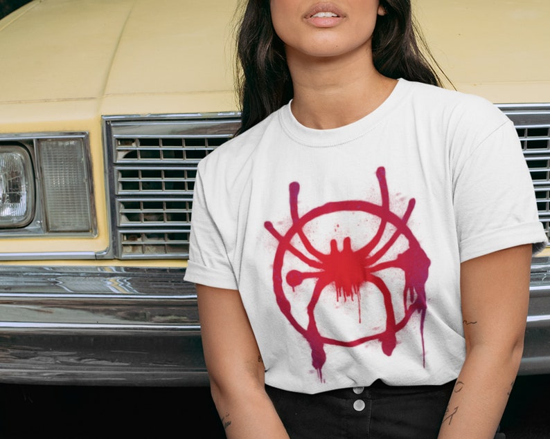 May include: White t-shirt with a red and pink spray paint design of a spider. The spider is in a circle with drips of paint coming from the circle.