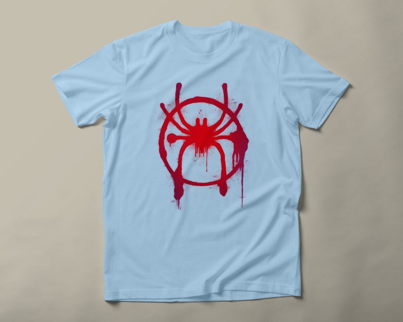 May include: Light blue t-shirt with a red and pink spray paint design of a spider in a circle.