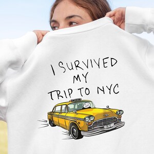 May include: White sweatshirt with the text "I survived my trip to NYC" in black handwritten font. A yellow taxi cab with a cartoon face is illustrated below the text.