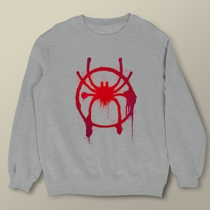 Puede incluir: Sudadera gris con un s&iacute;mbolo de Spider-Man rojo y rosa en la parte delantera. El s&iacute;mbolo es un c&iacute;rculo con una ara&ntilde;a dentro y tiene un efecto de pintura goteando.