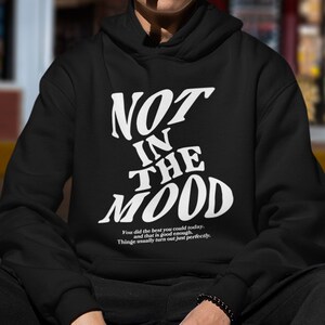 May include: Black hooded sweatshirt with white text that reads "NOT IN THE MOOD". The text is in a stylized font. Below the text is a smaller white text that reads "You did the best you could today. And that is good enough. Things usually turn out just perfectly."
