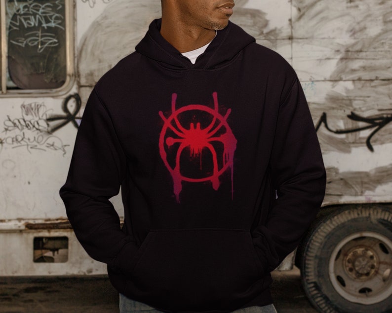 Puede incluir: Una sudadera con capucha negra con un s&iacute;mbolo de Spider-Man rojo y rosa en la parte delantera. El s&iacute;mbolo es una telara&ntilde;a estilizada con una ara&ntilde;a en el centro.