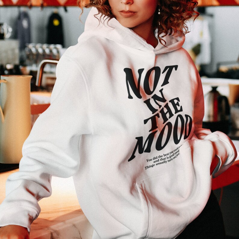 May include: White hoodie with black text that reads "NOT IN THE MOOD". The text is in a stylized font and is angled diagonally across the front of the hoodie. There is a small text block below the main text that reads "You did the best you could... and that is good enough. Things usually turn out just..."
