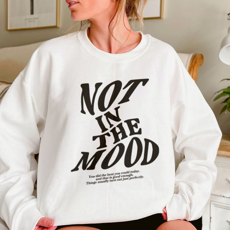 May include: White sweatshirt with black text that reads "NOT IN THE MOOD". The text is in a stylized font. Below the text is a smaller font that reads "You did the best you could today, and that is good enough. Things usually turn out just perfectly."