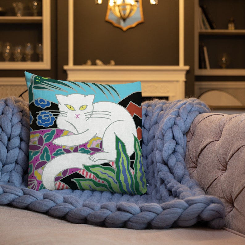 May include: A white cat with yellow eyes and a pink nose is depicted on a throw pillow. The cat is surrounded by colorful flowers and leaves on a blue background.