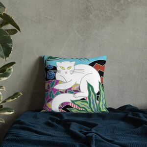 May include: A white cat with yellow eyes and a pink nose is lying on a colorful pillow. The pillow has a blue background with green, red, and purple flowers and leaves.