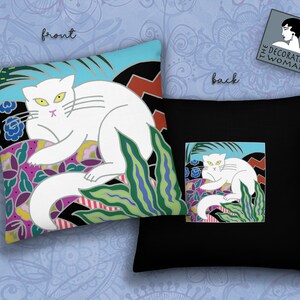 May include: A white cat with yellow eyes is lying on a colorful floral pattern. The cat is surrounded by green leaves and a blue sky. The pillow is black with a white cat on a colorful floral pattern. The pillow has the text "The Decorated Woman" on it.
