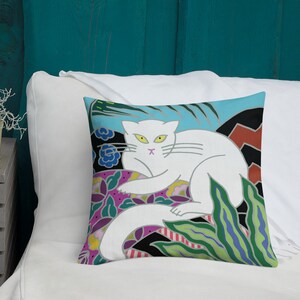 May include: A white cat with yellow eyes sits on a colorful floral patterned pillow. The pillow has a blue background with green, pink, and black accents.
