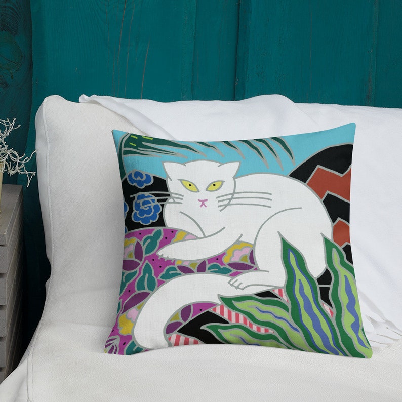 May include: A white cat with yellow eyes sits on a colorful floral patterned pillow. The pillow has a blue background with green, pink, and black accents.