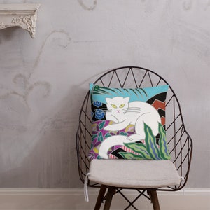 May include: A white cat with green eyes is lying on a colorful pillow with a floral and geometric design. The pillow is on a black metal chair with a white cushion.