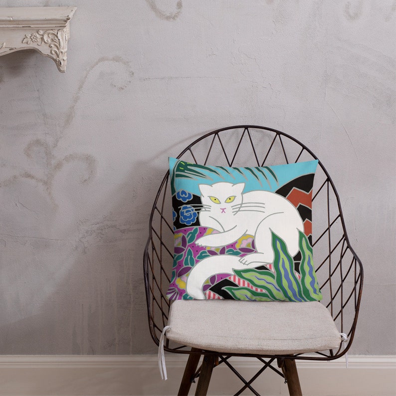 May include: A white cat with green eyes is lying on a colorful pillow with a floral and geometric design. The pillow is on a black metal chair with a white cushion.