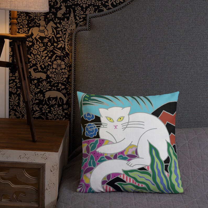 May include: A white cat with yellow eyes is lying on a colorful floral patterned pillow. The pillow has a blue background with green, purple, and red accents. The cat is surrounded by green leaves.