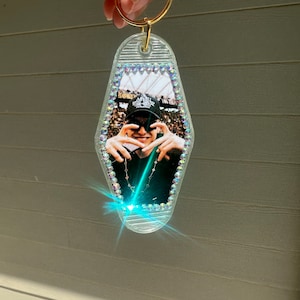 May include: A clear plastic motel key chain with a photo of a person wearing a black hat and making a heart shape with their hands. The keychain is decorated with iridescent rhinestones.