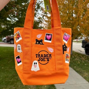 Peut inclure: Sac fourre-tout en toile orange vif avec le logo Trader Joe's. Le sac est orné d'autocollants sur le thème d'Halloween, dont des fantômes, des chats noirs, des cœurs roses et des feuilles d'automne. Il a deux poignées et une poche avant.