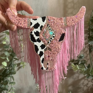 May include: Pink and black cow skull with glitter and fringe. The skull is painted with a black and white cow pattern on one side and pink glitter on the other side. The fringe is pink and hangs from the bottom of the skull.