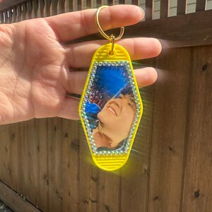 May include: Yellow motel key chain with a photo of a person inside. The photo is surrounded by rhinestones.