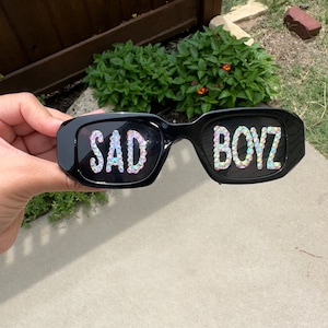 May include: Black sunglasses with the words "SAD BOYZ" spelled out in iridescent rhinestones.