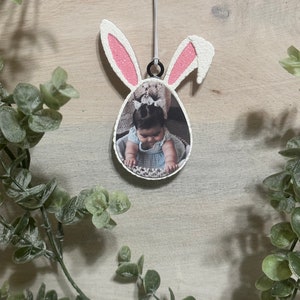 May include: Easter-themed photo ornament shaped like a bunny with pink ears and a photo of a baby. The ornament is hanging from a white string. The background is a light wood surface, and the ornament is surrounded by green foliage.