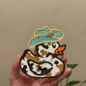 May include: A white and brown cow print rubber duck with a blue and gold glitter cowboy hat. The duck has a black outline and a yellow beak.