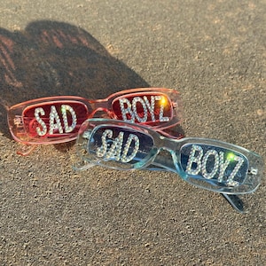 May include: Three pairs of sunglasses with rhinestone embellishments. The sunglasses are in pink, blue, and clear frames. The words "SAD" and "BOYZ" are spelled out in rhinestones on the sunglasses.
