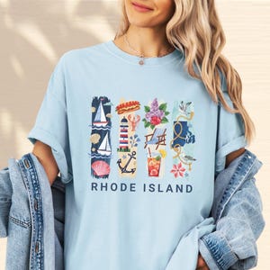 May include: Light blue t-shirt featuring a colorful graphic of Rhode Island icons, including a sailboat, lighthouse, and cocktail. The text "RHODE ISLAND" is printed in navy blue below the graphic. The shirt is a casual, comfortable style.