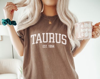 Puede incluir: Una camiseta marrón de gran tamaño con el texto "TAURUS EST. 1994" en blanco. La camiseta la lleva puesta una persona.
