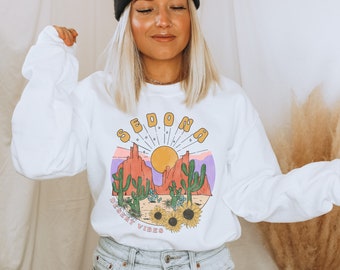 May include: White long-sleeve sweatshirt with a graphic print of a desert landscape with the text "SEDONA DESERT VIBES".