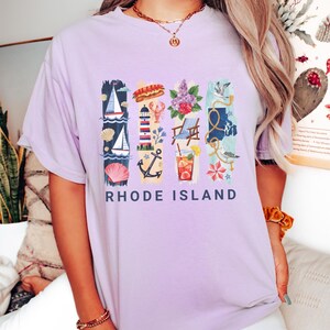May include: A light purple t-shirt with a colorful graphic design featuring nautical and summer-themed illustrations. The design includes a sailboat, lobster, lighthouse, and the text "RHODE ISLAND".