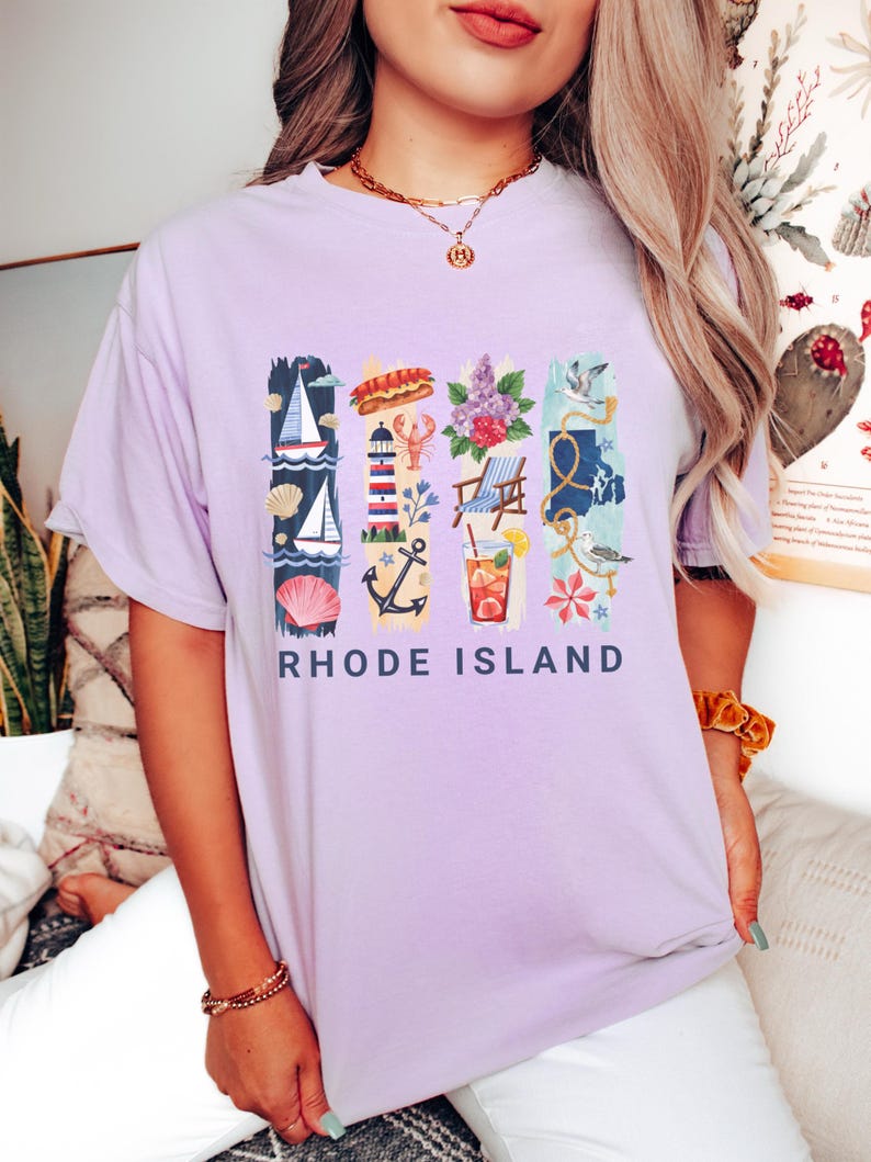 May include: A light purple t-shirt with a colorful graphic design featuring nautical and summer-themed illustrations. The design includes a sailboat, lobster, lighthouse, and the text "RHODE ISLAND".
