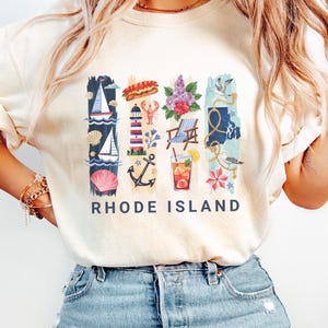 May include: Cream-colored t-shirt with a colorful Rhode Island graphic. The design includes nautical elements like sailboats, a lighthouse, and an anchor, along with a lobster, flowers, and a cocktail. The words "RHODE ISLAND" are printed in navy blue.
