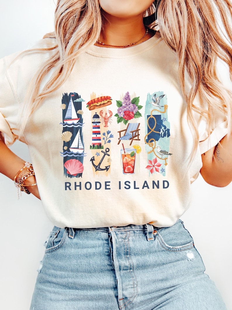 May include: Cream-colored t-shirt with a colorful Rhode Island graphic. The design includes nautical elements like sailboats, a lighthouse, and an anchor, along with a lobster, flowers, and a cocktail. The words "RHODE ISLAND" are printed in navy blue.
