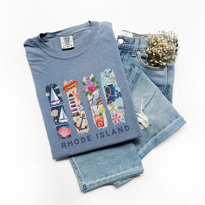 May include: A blue Comfort Colors t-shirt with a graphic of Rhode Island imagery, including a sailboat, lighthouse, flowers, and a cocktail. The shirt is paired with light-wash denim jeans.