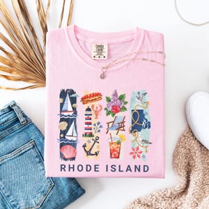 May include: A light pink t-shirt with a graphic design featuring Rhode Island imagery. The design includes a sailboat, lighthouse, lobster, flowers, a beach chair, and a map outline, with the words "RHODE ISLAND" at the bottom. A silver necklace is draped across the shirt.