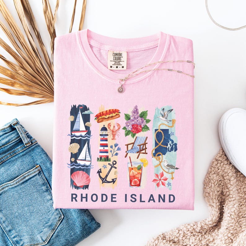 May include: A light pink t-shirt with a graphic design featuring Rhode Island imagery. The design includes a sailboat, lighthouse, lobster, flowers, a beach chair, and a map outline, with the words "RHODE ISLAND" at the bottom. A silver necklace is draped across the shirt.
