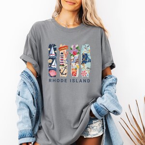 May include: Gray t-shirt with a colorful graphic featuring nautical and floral elements, including a sailboat, lighthouse, and flowers. The text "RHODE ISLAND" is printed below the design. The shirt has rolled-up sleeves.