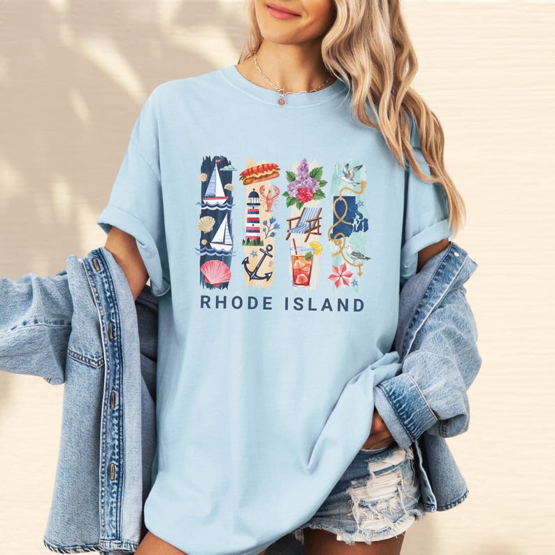 May include: Light blue t-shirt featuring a colorful graphic of Rhode Island icons, including a sailboat, lighthouse, and cocktail. The text "RHODE ISLAND" is printed in navy blue below the graphic. The shirt is a casual, comfortable style.