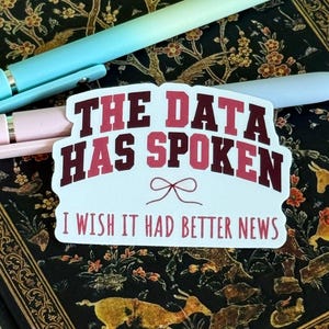 May include: A white die-cut sticker featuring the maroon text THE DATA HAS SPOKEN with a small bow icon and the text I WISH IT HAD BETTER NEWS below it. The sticker is placed on a black notebook with a gold floral and deer pattern, alongside two pens.