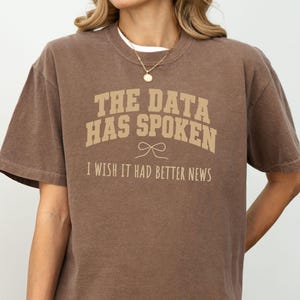 May include: A brown short-sleeve t-shirt featuring the beige text THE DATA HAS SPOKEN, a small line-art bow graphic, and the text I WISH IT HAD BETTER NEWS printed on the front. The shirt has a crew neckline and a relaxed, loose-fitting silhouette.