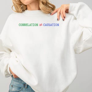 Op de afbeelding: Witte sweatshirt met de tekst "CORRELATION ≠ CAUSATION" in groen en blauw. De sweatshirt heeft een casual stijl, met lange mouwen en een ronde hals. De persoon draagt een blauwe jeans.