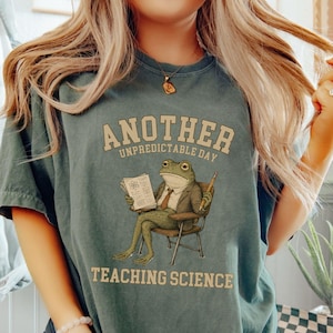 May include: A dark sage green t-shirt featuring a graphic of a frog wearing a suit and tie, sitting in a chair while holding a pencil and a document. The text reads ANOTHER UNPREDICTABLE DAY TEACHING SCIENCE in a tan, arched, block-style font.