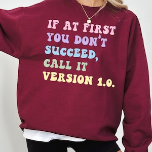 Könnte beinhalten: Ein weinrotes Sweatshirt mit dem Text "IF AT FIRST YOU DON'T SUCCEED, CALL IT VERSION 1.0." in Pastellfarben. Das Sweatshirt hat lange Ärmel und einen Rundhalsausschnitt. Die Person trägt schwarze Shorts und ein weißes Unterhemd.