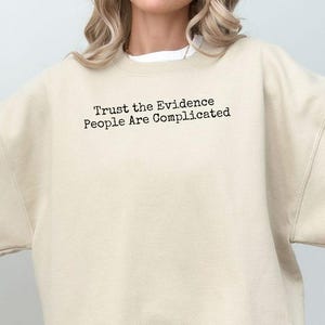 May include: A beige crewneck sweatshirt featuring the black text Trust the Evidence People Are Complicated printed on the upper chest. The garment has long sleeves, a ribbed neckline, and a relaxed fit, providing a neutral and simple appearance.