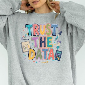 May include: A heather gray crewneck sweatshirt featuring the colorful text TRUST THE DATA in a crayon-like font. The design includes illustrations of a graph, a notebook, a test tube, and a calculator, all rendered in a vibrant, hand-drawn style.
