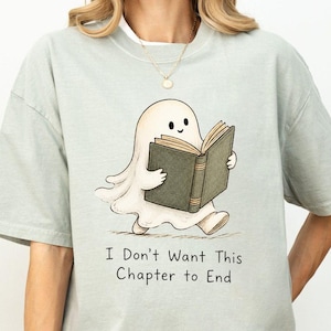 Könnte beinhalten: Ein helles salbeigrünes T-Shirt mit kurzen Ärmeln und einer Grafik eines kleinen weißen Geistes, der in einem aufgeschlagenen dunkelgrünen Buch liest. Unter dem Geist steht der Text I Don't Want This Chapter to End in einer einfachen schwarzen Schrift.