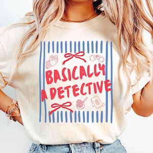 May include: A cream-coloured t-shirt featuring a blue vertical striped background with red line art illustrations of handcuffs, a magnifying glass, and bows. The text "BASICALLY A DETECTIVE" is printed in red across the centre of the design.