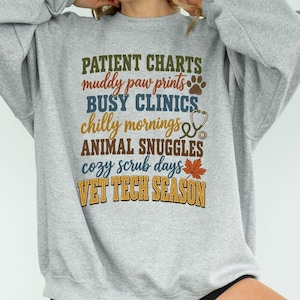 Puede incluir: Una sudadera gris claro de cuello redondo con texto multicolor que dice: PATIENT CHARTS, muddy paw prints, BUSY CLINICS, chilly mornings, ANIMAL SNUGGLES, cozy scrub days y VET TECH SEASON, junto a un estetoscopio y una hoja.