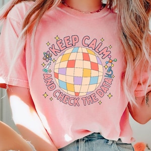 Puede incluir: Una camiseta de manga corta de color rosa claro con un gráfico central de una bola de discoteca multicolor rodeada por el texto "KEEP CALM AND CHECK THE DATA". El diseño incluye pequeños iconos científicos como un átomo, un vaso de precipitados y un microscopio.
