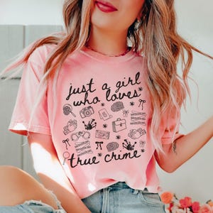 以下が含まれることがあります： 「just a girl who loves true crime」の黒い文字がプリントされた、薄いピンク色のTシャツ。虫眼鏡、カメラ、手錠など、犯罪に関連する様々なイラストが描かれています。