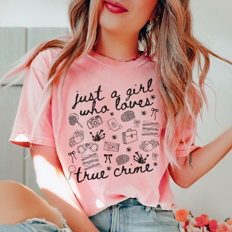 以下が含まれることがあります： 「just a girl who loves true crime」の黒い文字がプリントされた、薄いピンク色のTシャツ。虫眼鏡、カメラ、手錠など、犯罪に関連する様々なイラストが描かれています。