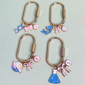 May include: Four gold-colored keychains with science-themed charms. Each keychain has a different charm, including a DNA strand, microscope, chemistry globe, and a flask. Each charm is paired with a pink bow and a letter.